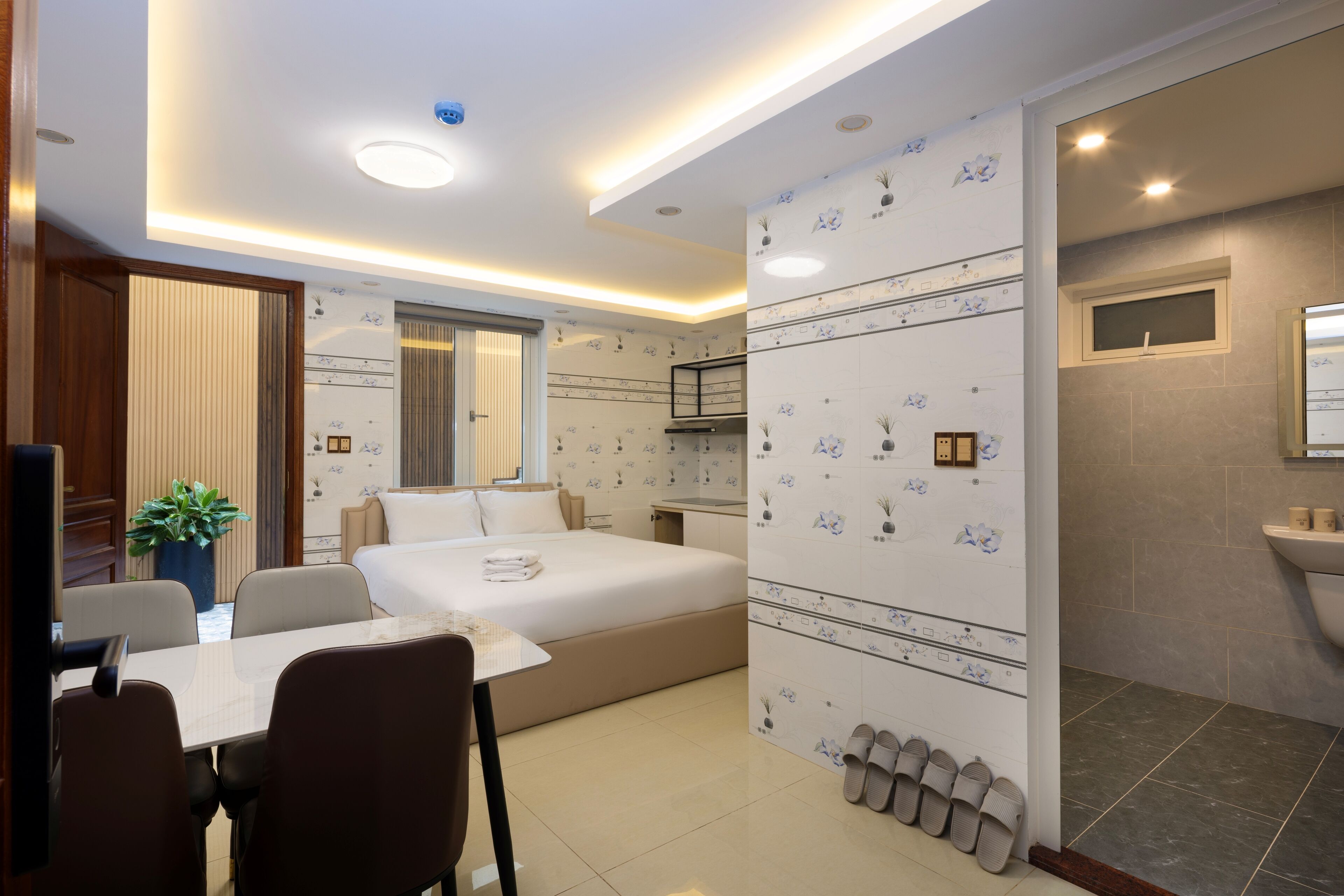 A modern hotel room with a bed, a dining table, chairs, a small kitchen area, and a bathroom with a sink and mirror.