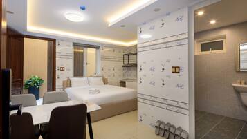 A modern hotel room with a bed, a dining table, chairs, a small kitchen area, and a bathroom with a sink and mirror.