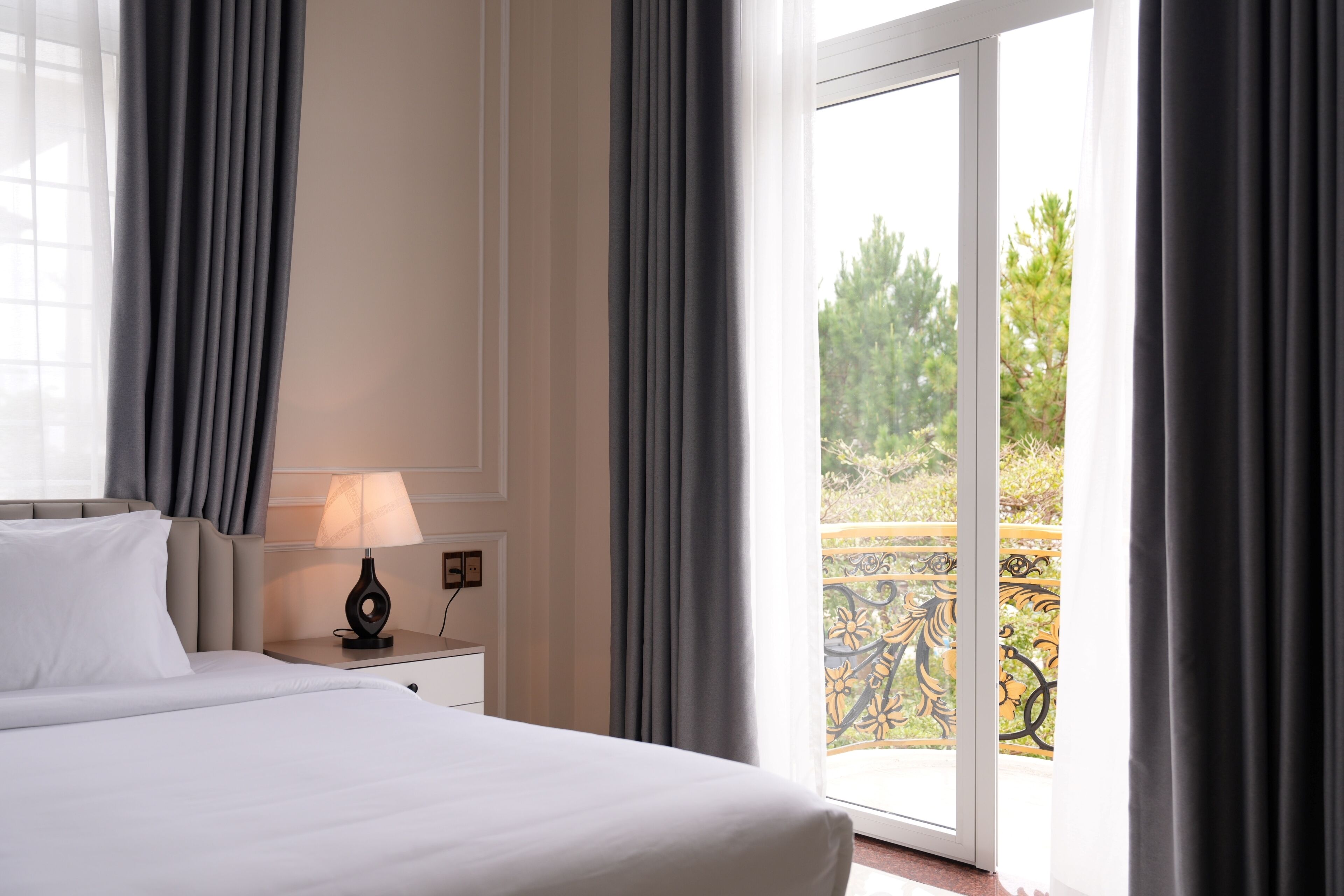 Una habitación de hotel con una cama, una mesita de noche con lámpara y vistas a un balcón con mobiliario exterior.