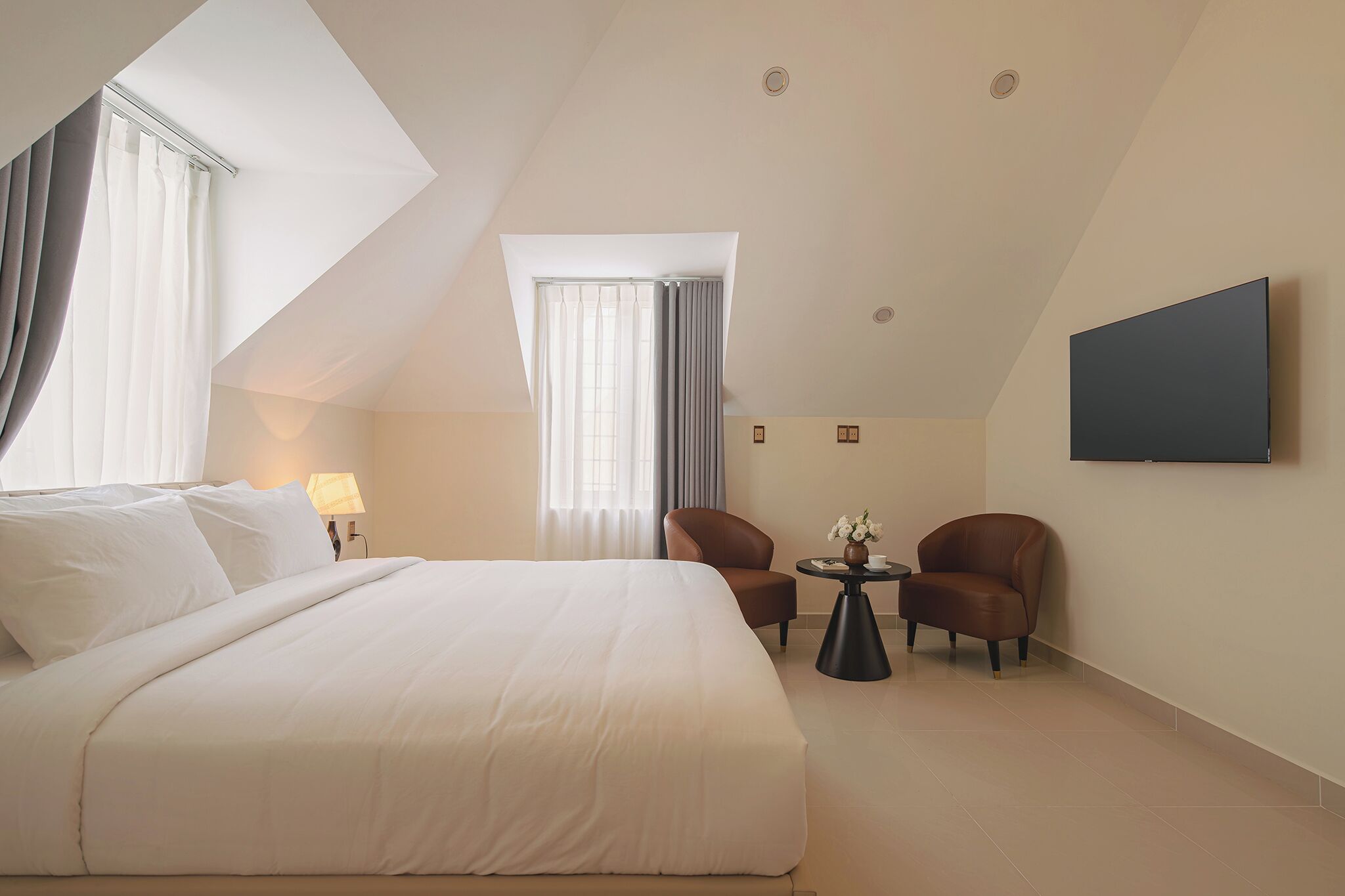 A hotel room with a large bed, two leather chairs, a small table with flowers, a flat-screen TV, and a skylight.
