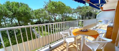 A balcony with white plastic chairs and a table, a blue awning, and a view of trees.