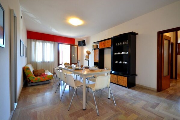 A dining area with a table set for four, surrounded by white chairs. A sofa with colorful cushions is against the wall. A black cabinet with glass doors is to the right.