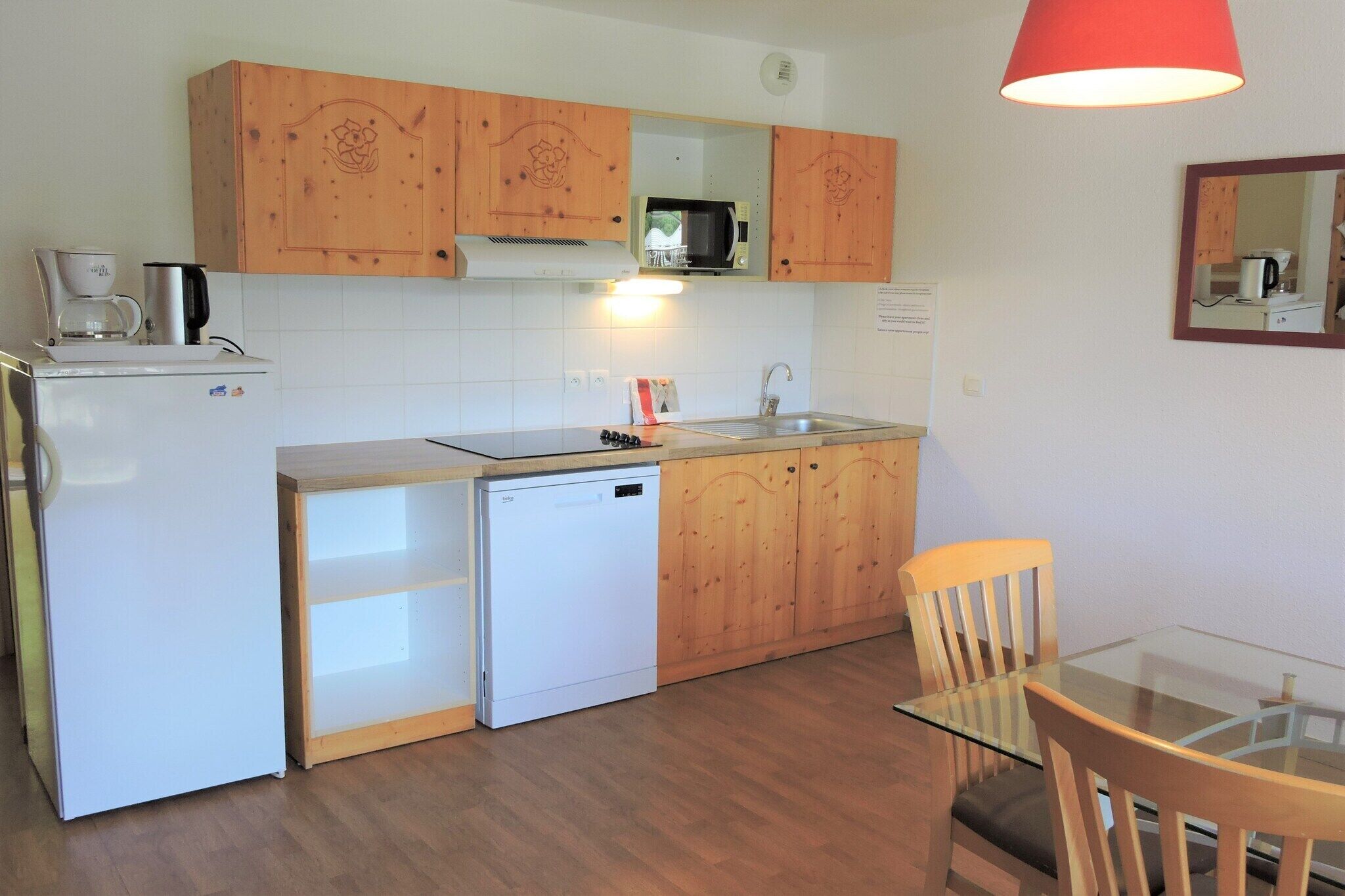 Een kleine keuken met houten kastjes, een magnetron, een spoelbak en een glazen tafel met een stoel.