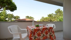 Un balcón con una mesa cubierta con mantel floreado, dos sillas blancas de plástico y un vaso de agua.