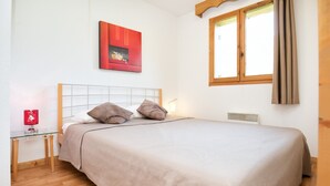 En snyggt bäddad säng med ett beige överkast och två kuddar, ett litet sängbord med en röd lampa, ett fönster med träramar och beige gardiner samt en röd, abstrakt tavla på väggen.