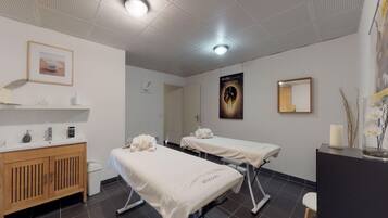 Une salle de spa propre et moderne, équipée de deux tables de massage, d’un meuble en bois, d’un lavabo et d’un miroir mural.