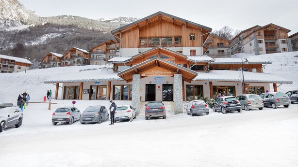 A multi-story wooden building with a stone base, labeled "Residence Le Hameaux des Eaux d'Olle," surrounded by parked cars and snow-covered ground.