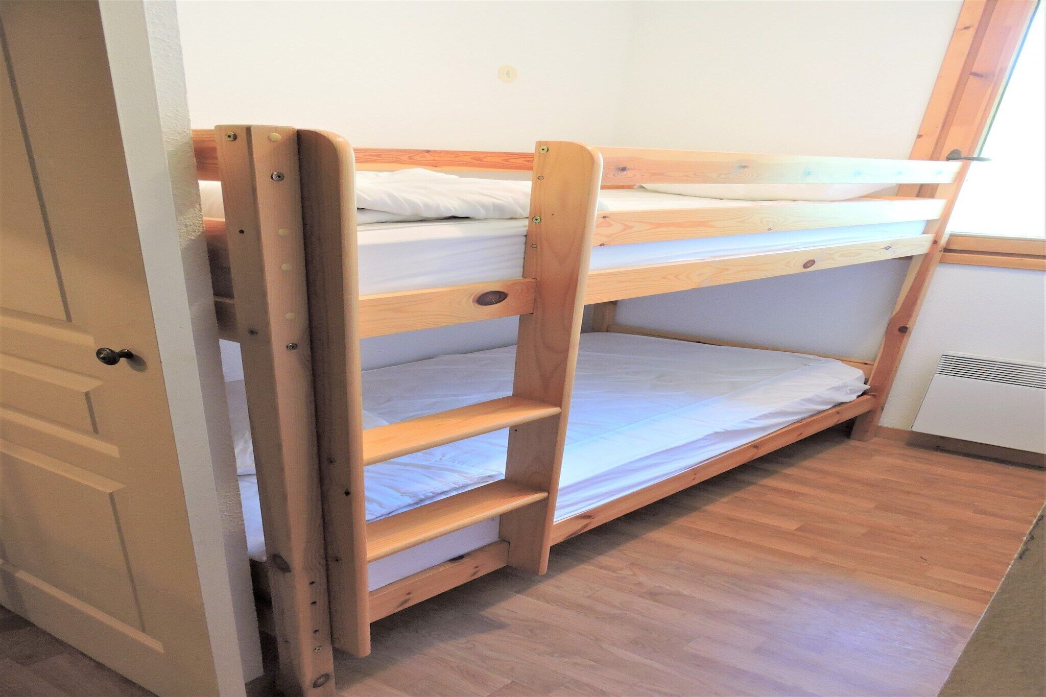 Un lit superposé avec une structure en bois et du linge de lit blanc, une porte sur la gauche et un petit appareil électroménager blanc sur la droite.