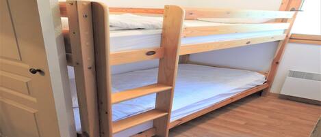 Un lit superposé avec une structure en bois et du linge de lit blanc, une porte sur la gauche et un petit appareil électroménager blanc sur la droite.