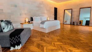 A modern hotel room with a wooden herringbone floor, a grey sofa, a white bed, and two bedside tables with lamps.