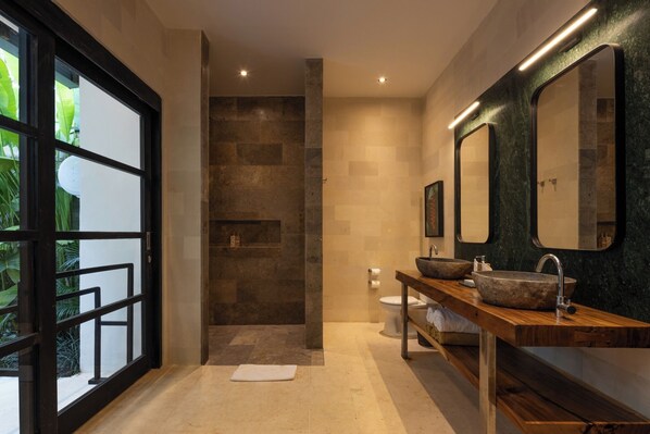 Une salle de bain moderne dotée d’un grand miroir, de deux vasques en pierre, d’une zone de douche et d’une vue sur la verdure grâce à une porte en verre.