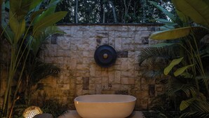A large, oval-shaped bathtub in a natural setting with a stone wall and lush greenery.