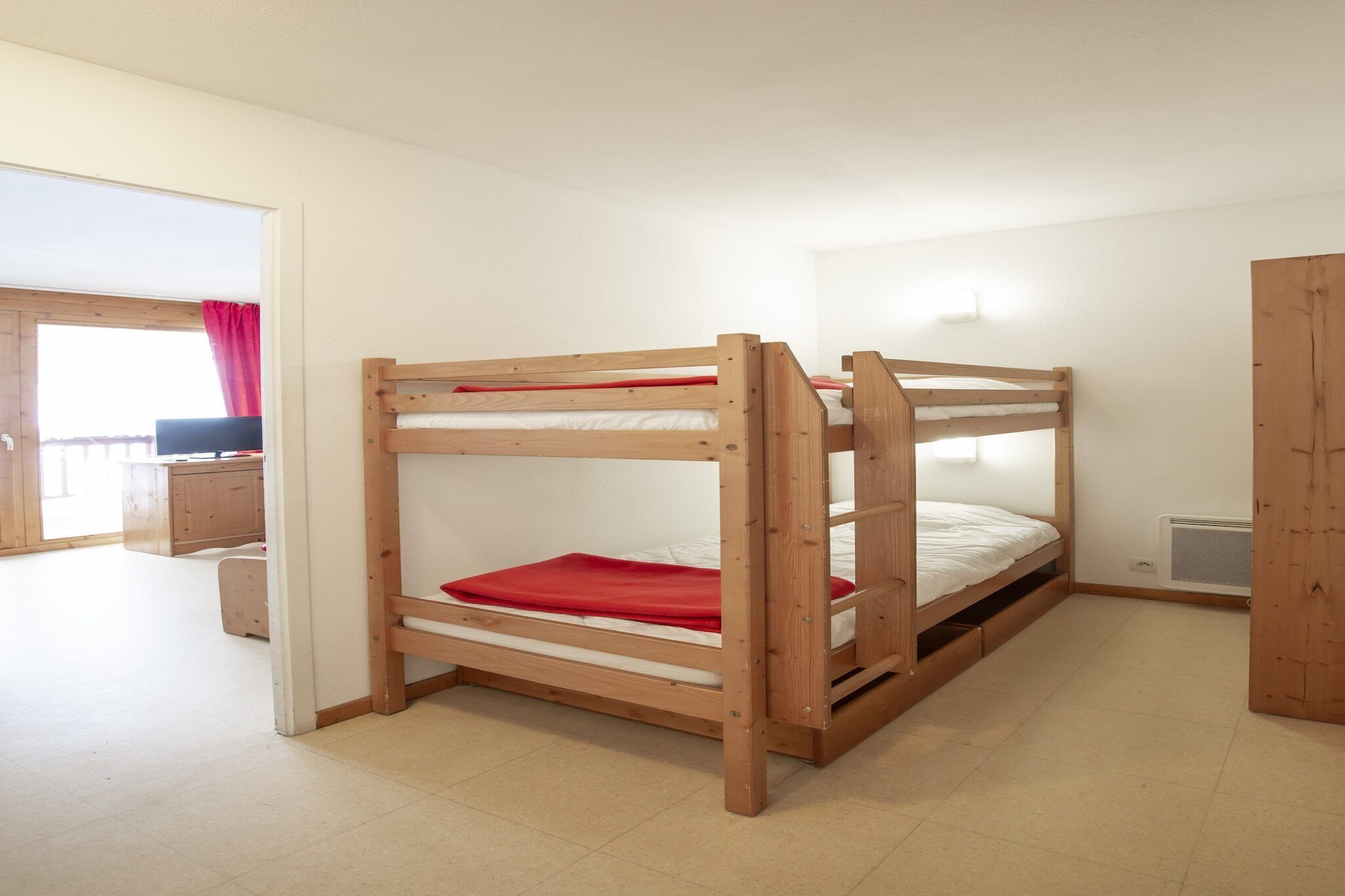 A bunk bed room with a wooden frame, a red blanket, and a white mattress. There is a window with a red curtain and a door leading to another room.
