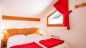 A bedroom with a wooden headboard, two beds with white bedding and red blankets, a nightstand with a lamp, a window with red curtains, and a wooden shelf with two round objects. - Apartment in Val Cenis near River (Val Cenis)