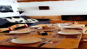 Une table à manger dressée avec des assiettes, des couverts et des verres, un banc en bois, un canapé agrémenté d’un coussin à motifs noir et blanc, et une cheminée en arrière-plan.
