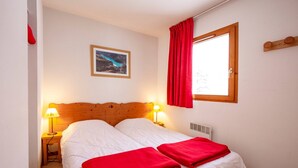 Una cama doble con ropa de cama blanca y mantas rojas, dos lámparas de noche, un cabecero de madera, una ventana con cortinas rojas y un cuadro enmarcado en la pared.