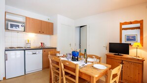 Una cocina compacta con armarios de madera, microondas, lavavajillas, una mesa de comedor con sillas, televisión y un mueble pequeño con lámpara.