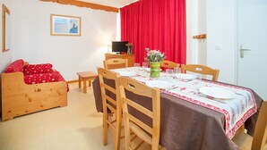 Ett mysigt rum med en säng i trä, ett matbord dukat med tallrikar och glas, en tv-apparat på ett träskåp och en röd gardin.