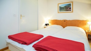 A double bed with white bedding and two red blankets, a wooden headboard, a nightstand with a lamp, and a framed picture on the wall. - Apartment with 2 rooms, balcony (Val Cenis)