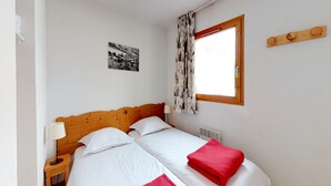 Cama doble con ropa de cama blanca y roja, cabecera de madera, mesita de noche con lámpara, ventana con cortinas y perchero en la pared.