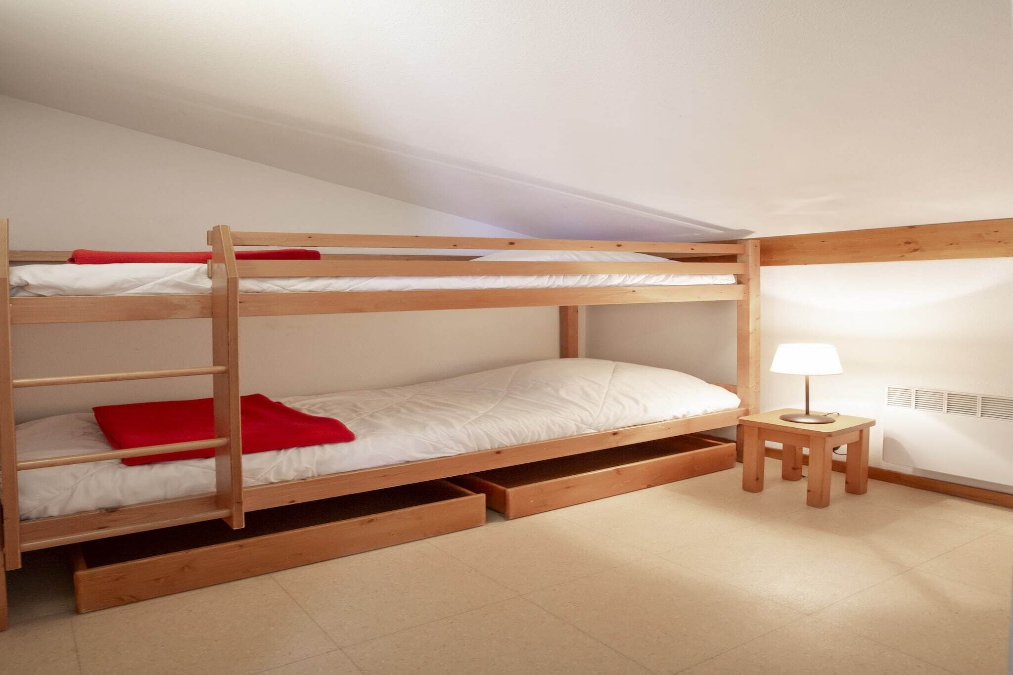 Een stapelbed met opbergladen, een tafeltje met een lamp en een radiator aan de muur.