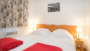 Een net opgemaakt bed met witte en rode lakens, twee bedlampjes, een houten hoofdbord, een aan de muur hangend schilderij en een raam met gordijnen.