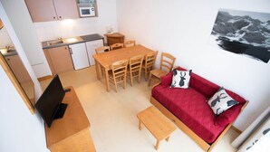 Een knus woongedeelte met een kitchenette, een eettafel, een rode bank en een afbeelding van een berglandschap.