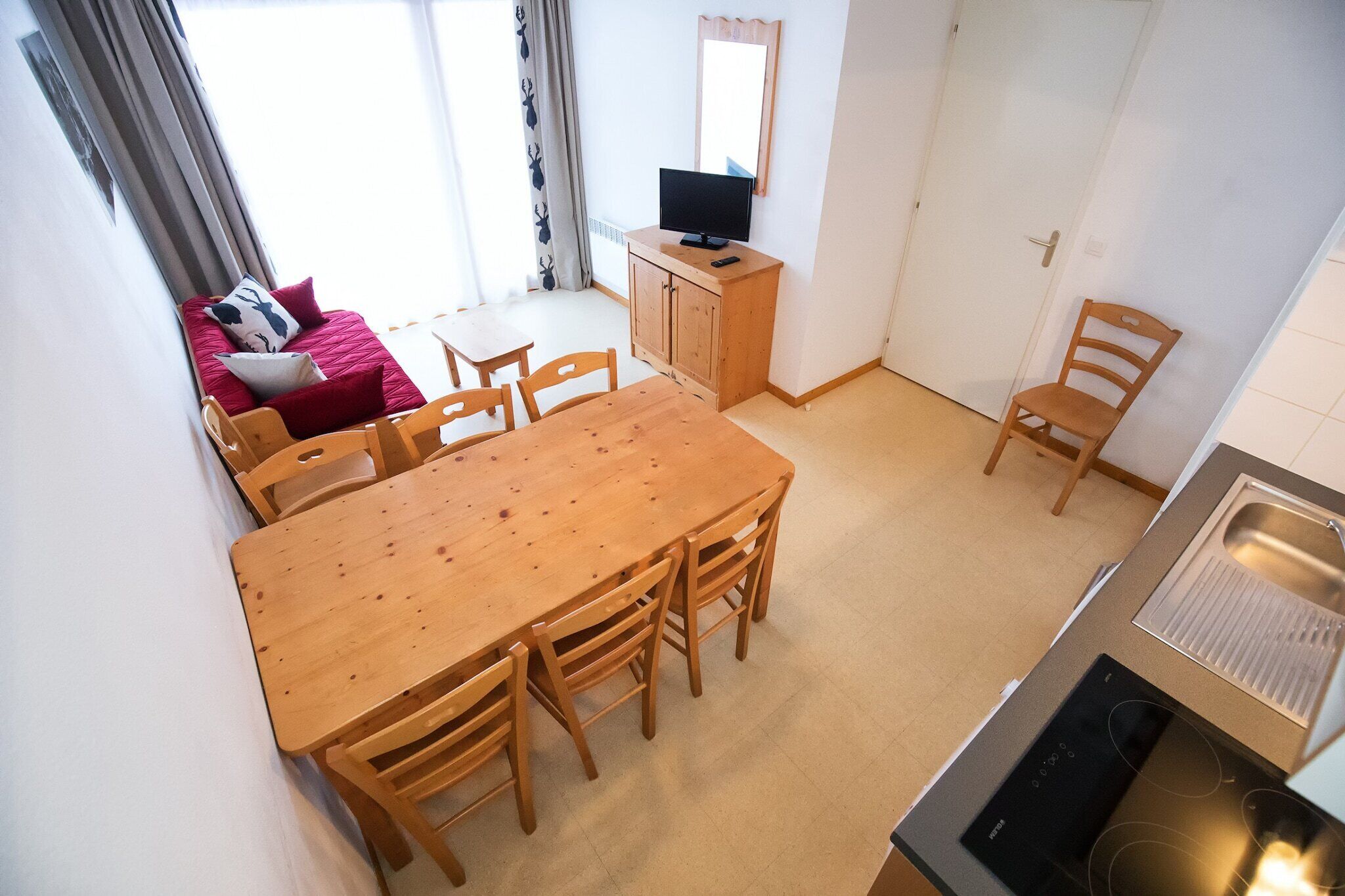 Ett kompakt bostadsutrymme med en matplats, ett litet kök och en liten sittgrupp med en röd soffa och kuddar.