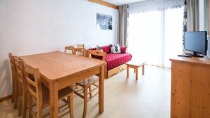 Ein Zimmer mit einem Holzespeise Tisch und Stühlen, einem Sofa mit roten Kissen, einem Fernseher auf einem Holzschrank und einem Fenster mit Vorhängen.