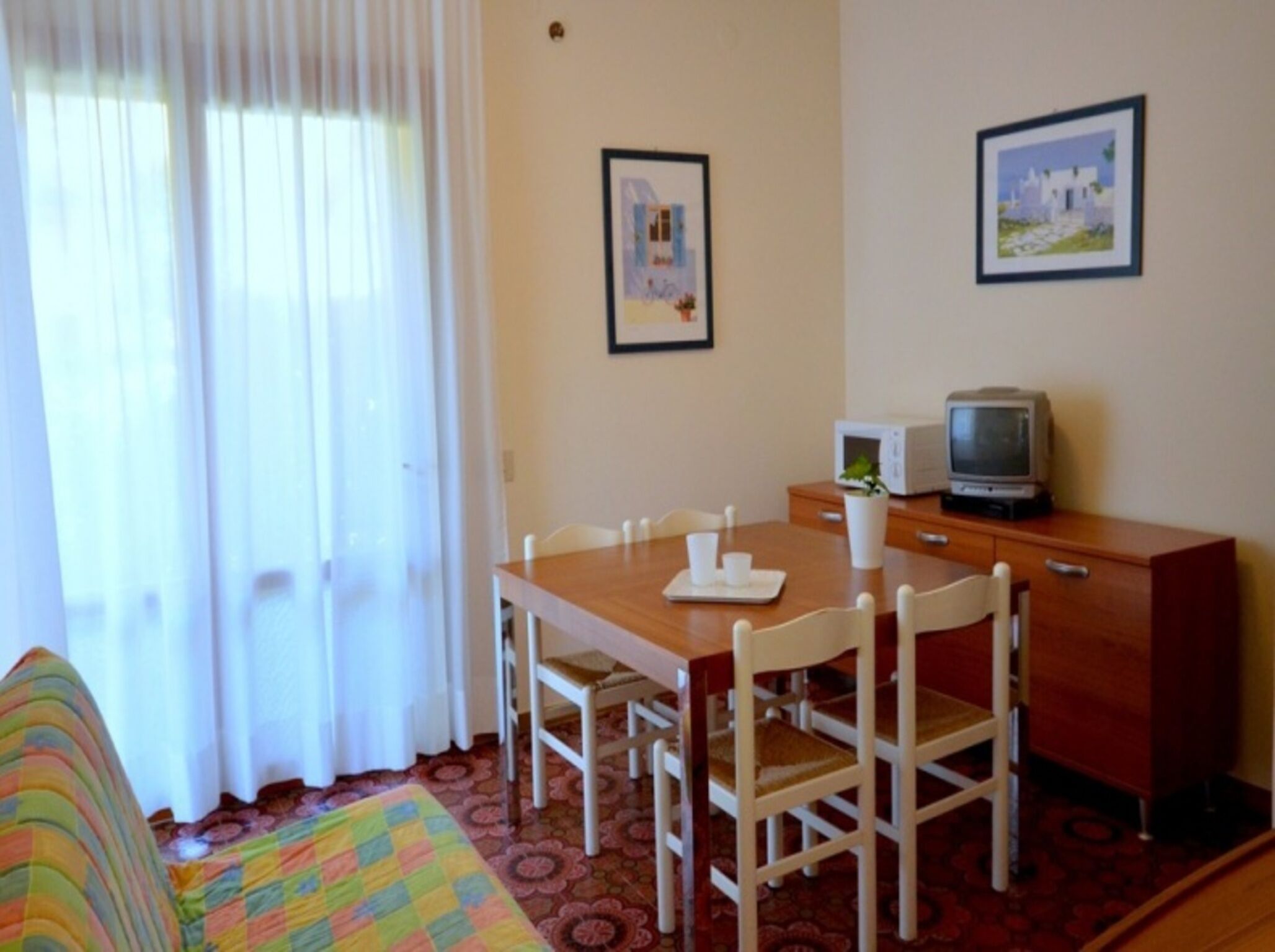 Habitación de hotel con una mesa de comedor de madera, sillas blancas, un televisor pequeño sobre un mueble de madera y dos cuadros en la pared.
