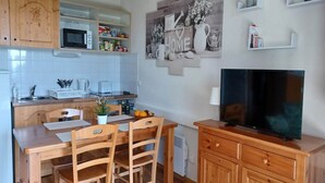 Een kleine keuken met houten kastjes, een magnetron, een eettafel met stoelen, een televisie op een houten standaard en een wandplank met decoratieve objecten.