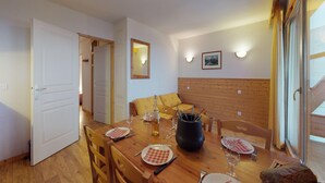 Un comedor con una mesa de madera dispuesta para dos personas, una olla negra, copas de vino y una botella. Hay un banco de madera y una puerta que da a otra habitación.