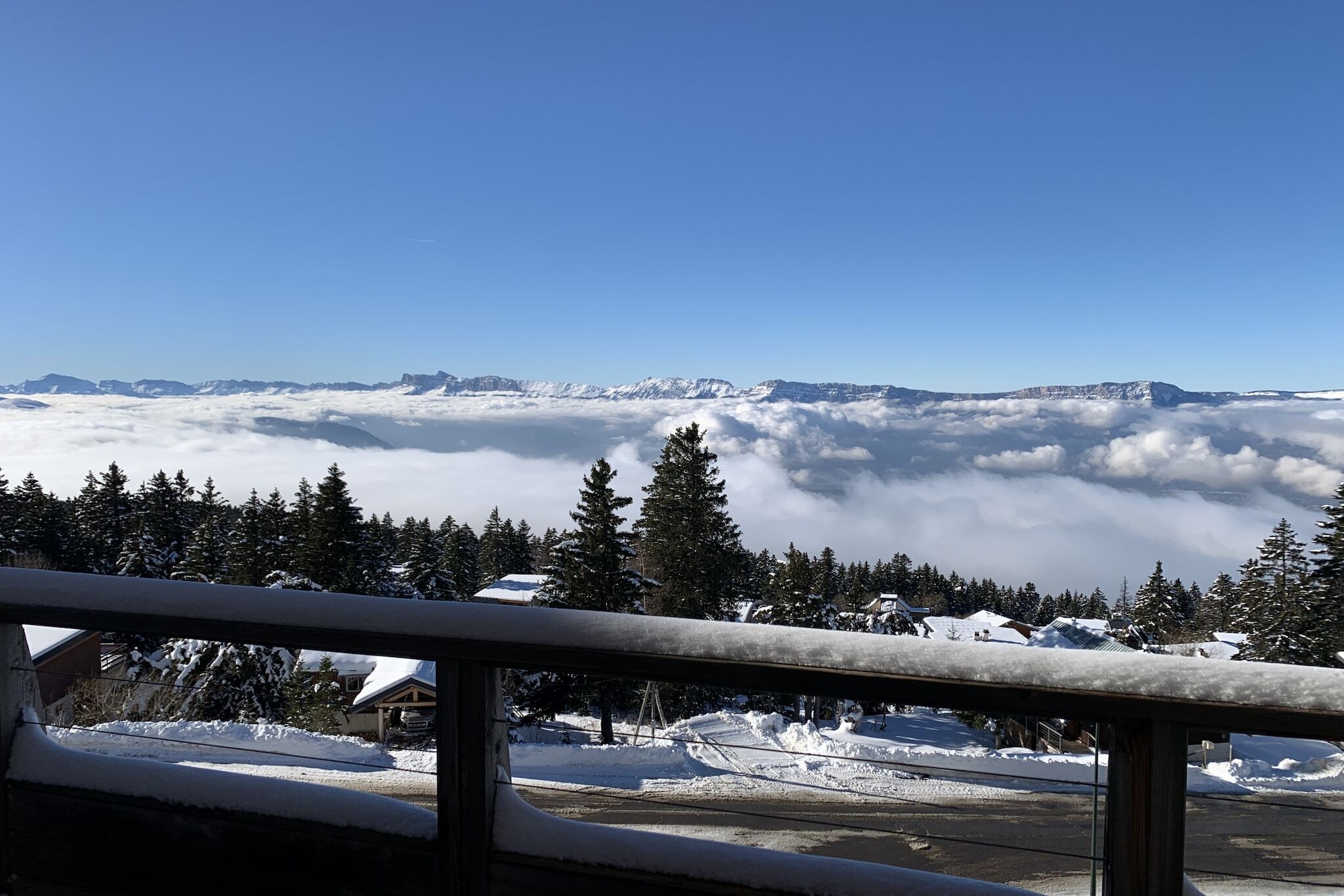 Un paysage montagneux enneigé, avec un ciel dégagé, une zone boisée et un bâtiment dont les toits sont recouverts de neige.