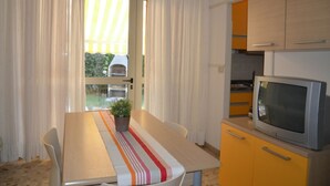 A compact kitchen with a dining area, featuring a table with a striped tablecloth, a potted plant, and a television on a cabinet. - Holiday flat with barbecue and private garden (Bibione Pineda)