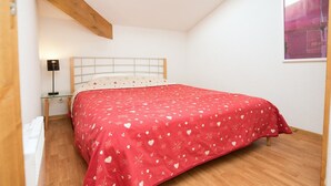 A bedroom with a wooden floor, a bed with a red bedspread, a nightstand with a lamp, and a white headboard.