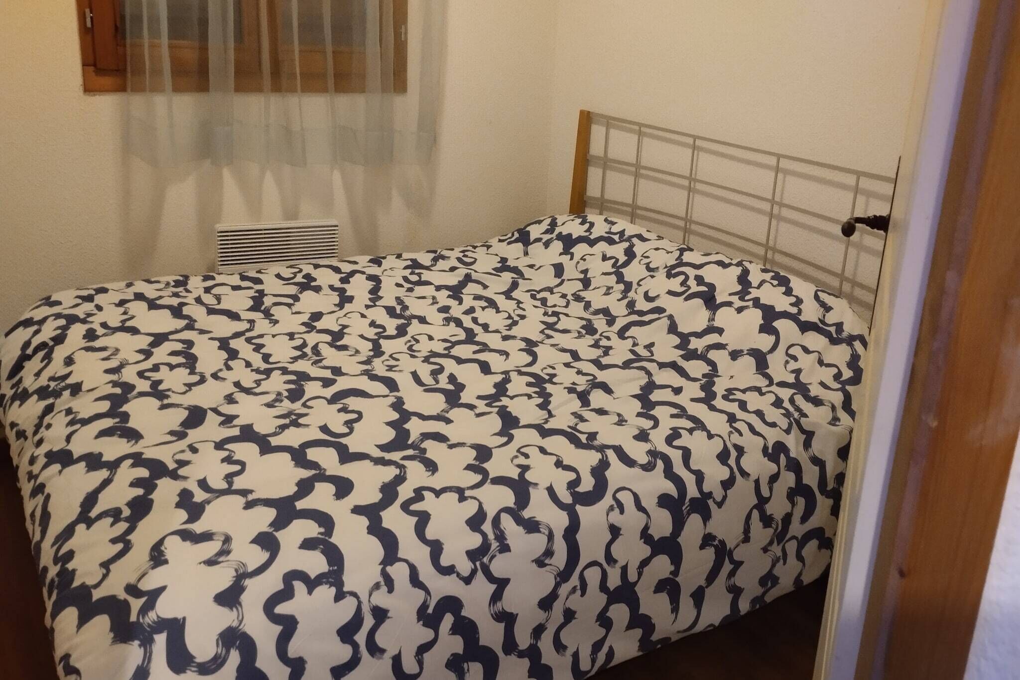Un lit simple avec une couvre-lit à motifs, une fenêtre avec des rideaux légers et un radiateur fixé au mur.
