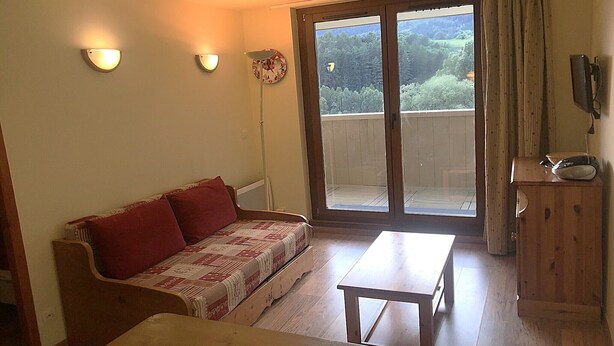 Una habitación con sofá de madera, una mesita, un armario de madera y un ventanal corredero que da a un balcón con vistas a zonas verdes.