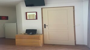 Una habitación con puerta cerrada, un pequeño aparador de madera con una computadora portátil, un televisor montado en la pared y un refrigerador en la esquina.
