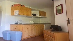 Cocina compacta con gabinetes de madera, microondas, cafetera y teléfono sobre una encimera. Hay dos cubos de basura y un mueble pequeño con cajones.
