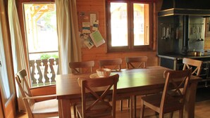 A wooden dining table with six chairs, a kitchen area with a stove, and a window with curtains.