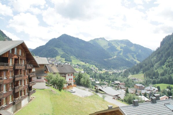 A mountain lodge with a wooden facade, multiple balconies, and a view of a valley with houses and mountains in the background.