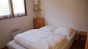 A bedroom with a bed, a bedside table, a lamp, a heater, and a window with curtains. - Jonquilles apartment for 4 people (Châtel)