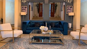 Una sala de estar con un sofá azul, dos sillones blancos, una mesa de centro de vidrio y una araña.