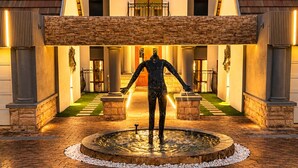 Un edificio con entrada central, paneles solares, fachada de piedra y una fuente con una estatua.