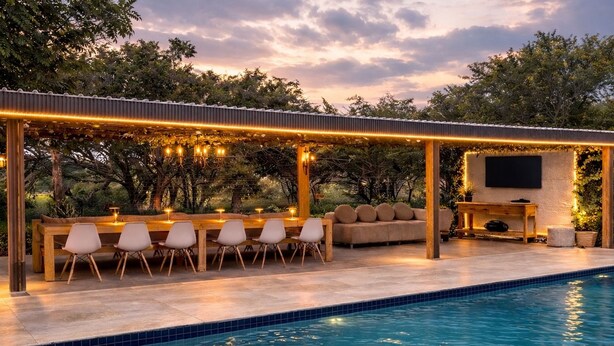 Zona junto a la piscina con una terraza de madera, espacio de estar con sofá y sillas, y un televisor fijado a la pared.