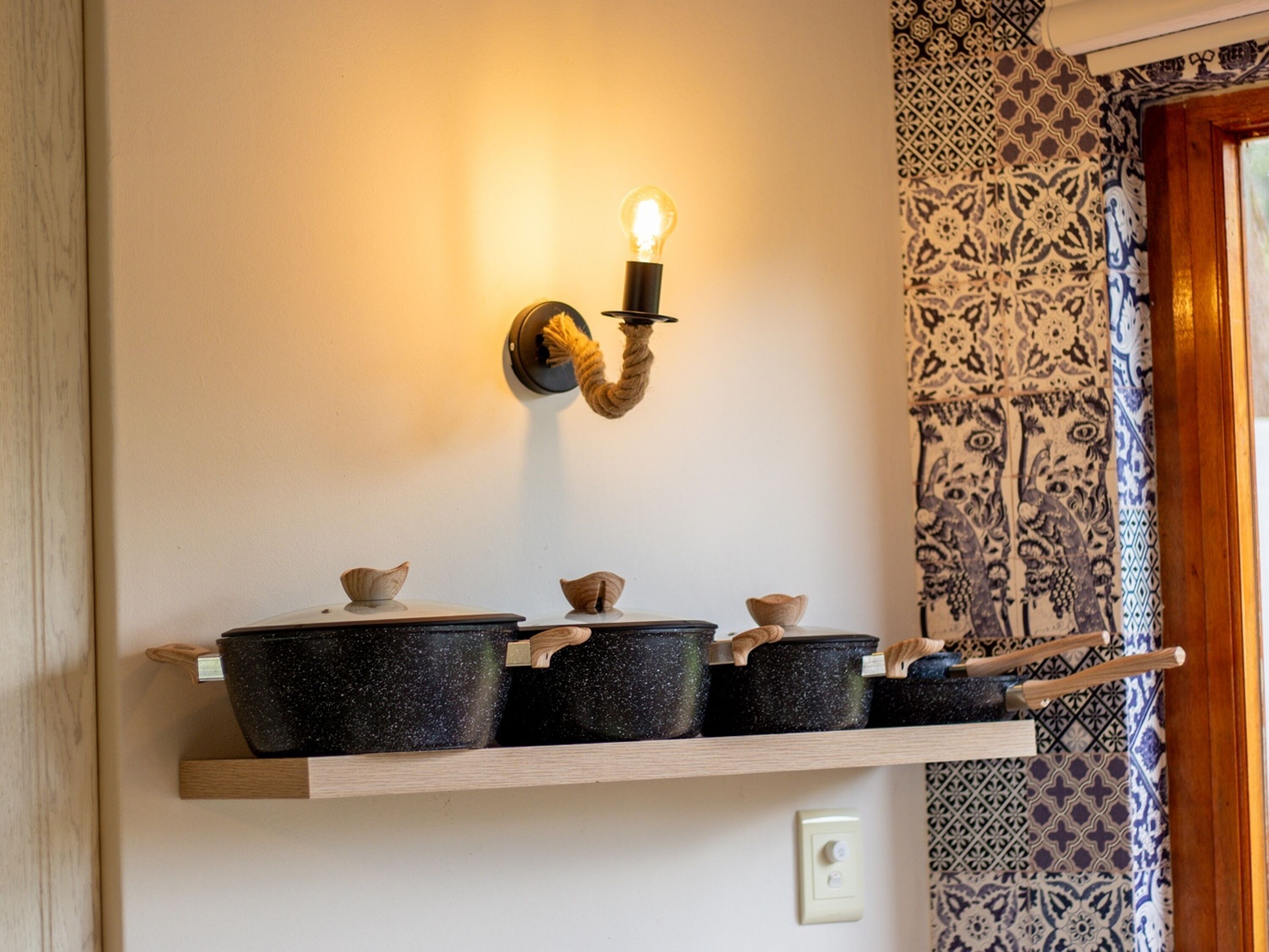 Une cuisine avec une étagère en bois sur laquelle sont posées plusieurs casseroles noires, un luminaire fixé au mur et un papier peint à motifs.