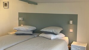 Quarto com duas camas, cada uma com várias almofadas e cabeceira. Há luminárias de cabeceira e uma pequena mesa de apoio.