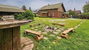 Un edificio de madera rústico con techo verde, rodeado de un patio cubierto de césped con mesas de picnic y una fogata de piedra.