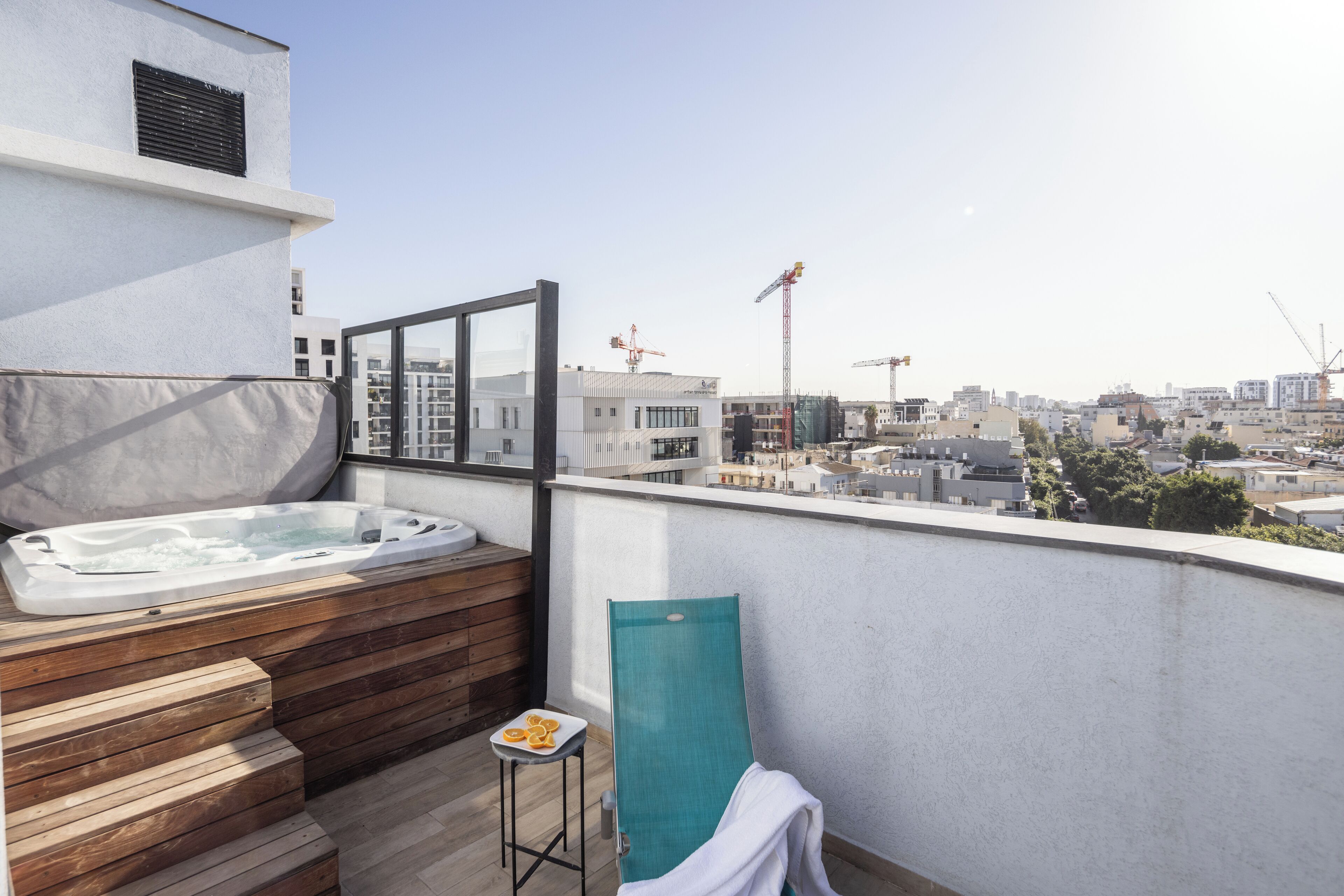 A rooftop terrace with a hot tub, wooden steps, a small table with oranges, and a view of the cityscape and construction cranes.