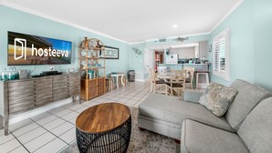 A modern living room with a sofa, coffee table, and TV. There is a kitchen area with cabinets and a dining table. - Clearwater 2A (Gulf Shores)